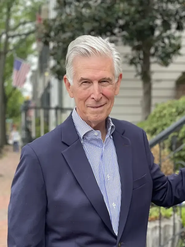 Man with gray hair in a navy blazer and striped shirt standing on an urban street lined with trees and historic buildings, hand resting on a railing.