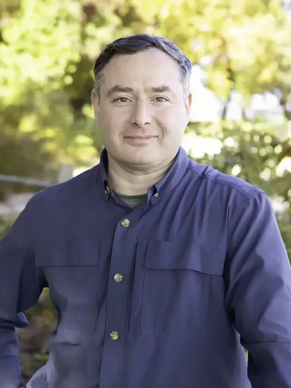 Man with dark hair wearing a navy button-up shirt with chest pockets, photographed outdoors against a softly blurred background of green trees.